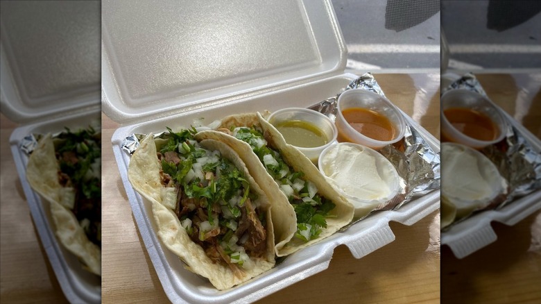 Two soft tacos on the left side of a foil-lined rectangular styrofoam to-go-box filled with meat, chopped onions, and cilantro; three clear plastic ramekins are on the right — one contains red sauce, one sour cream, and the third salsa verde
