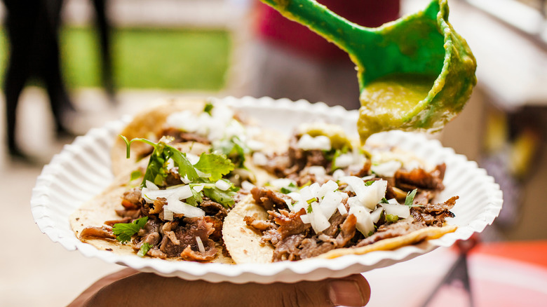 A hand holding a white paper plate with street tacos on it with a green ladle pouring verde sauce on top of them
