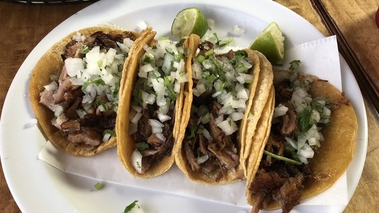 Four tacos in corn tortillas on a round white plate; each taco is filled with meat, chopped onions, and cilantro; two squeezed lime wedges are situated at the top