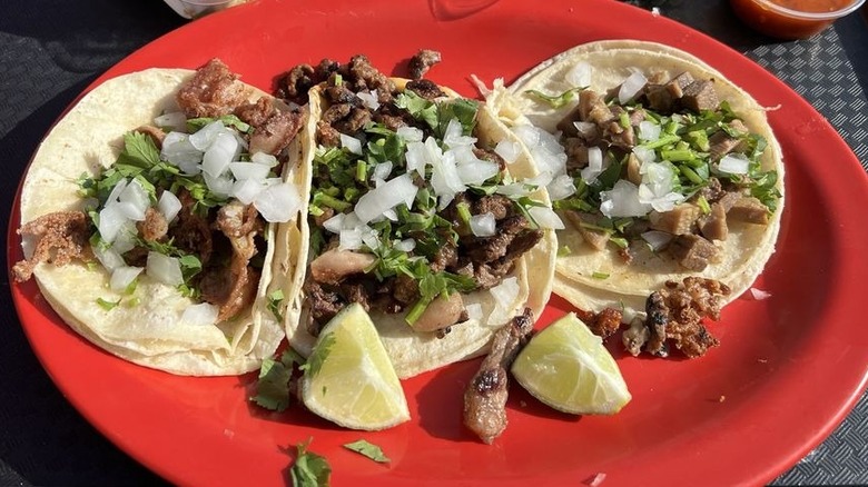 Three soft tacos in corn tortillas lined up on a red plate; each is filled with meat, chopped white onions, and cilantro. Two lime wedges sit at the bottom of the plate