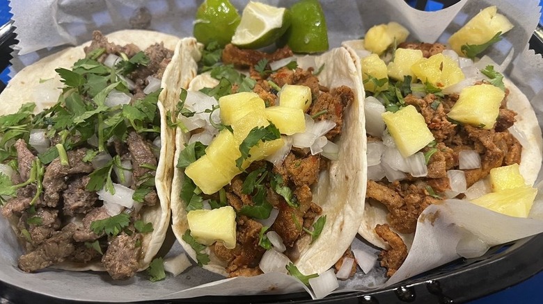 Close-up of three tacos in flour tortillas lined up on a plate: The left is filled with meat, chopped onions, and cilantrol; the other two are filled with meat, chopped onions, pineapple chunks and cilantro