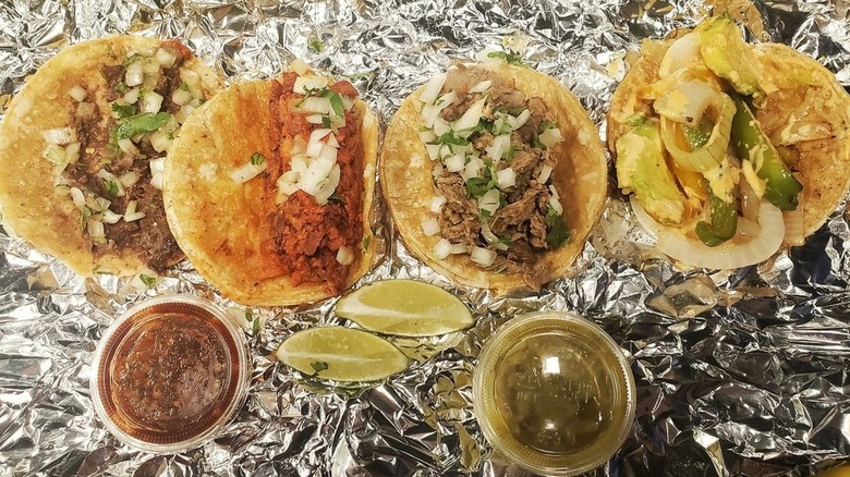 Four tacos on corn tortillas lined up on a piece of crumpled aluminum foil; the three tacos on the left are filled with meat, chopped white onions, and cilantro; the one on the right is filled with onions, green peppers, and avocado; clear plastic ramekins of red sauce, green sauce, and lime wedges sit below the tacos