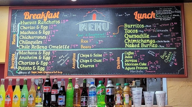 A colorful, handwritten chalk menu board with sections for breakfast, lunch, drinks, and appetizers; a shelf sits below the board that is lined with Mexican and American soda and drink bottles
