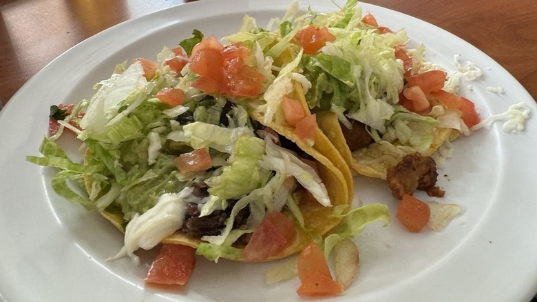 Two tacos in corn tortillas on a round white plate stuffed with meat, lettuce, sour cream, cotija cheese, and chopped tomatoes