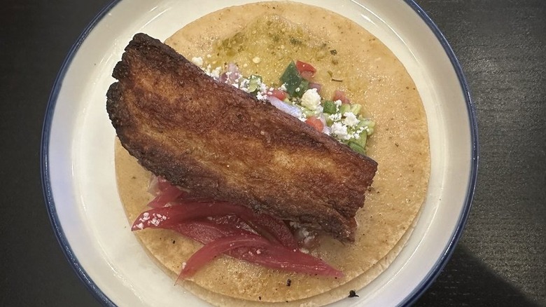A single pork belly taco on a corn tortilla iwth pickled onions, cotija cheese, and pico de gallo