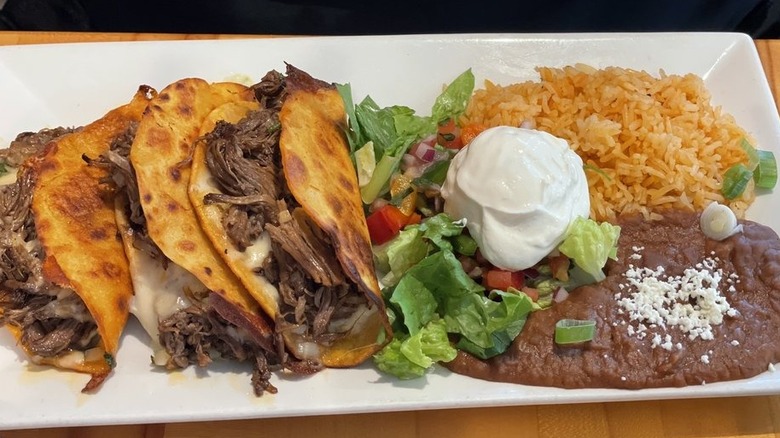 Three quesaberria tacos on the left side of a white rectangular plate; there's torn lettuce, chopped red onions, and tomoatoes in the center, a dollop of sour cream, and rice and beans on the right