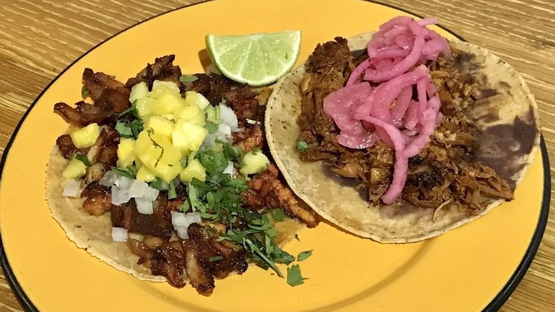 Two tacos on corn tortillas on a round yellow plate; the right one has meat, chopped onions, cilantro, and chopped pineapple; the right one has meat and pickled red onions; a lime wedge at the top of the plate