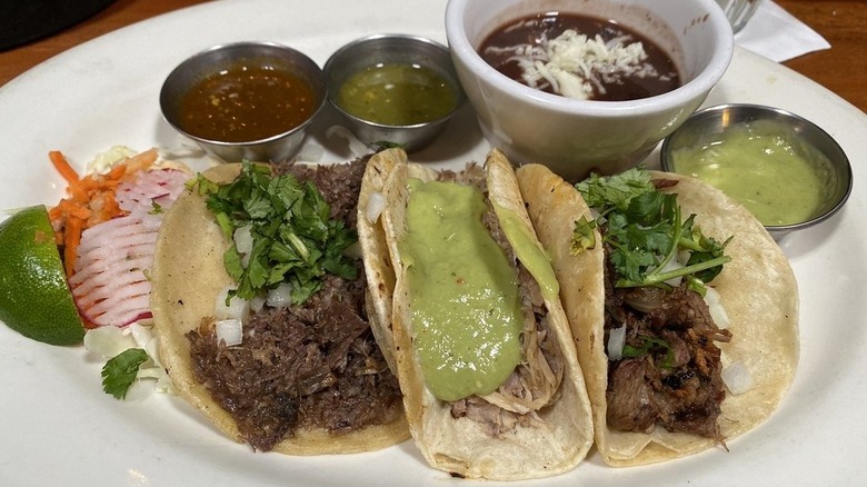 Three tacos on a round white plate; all three are filled with meat, the right is topped with chopped onions and cilantro, the middle with a green sauce, the left with chopped onions and cilantro; there's a lime wedge and julienne radish and carrots on the far right, and ramekins of red, green, and mole sauce line the top