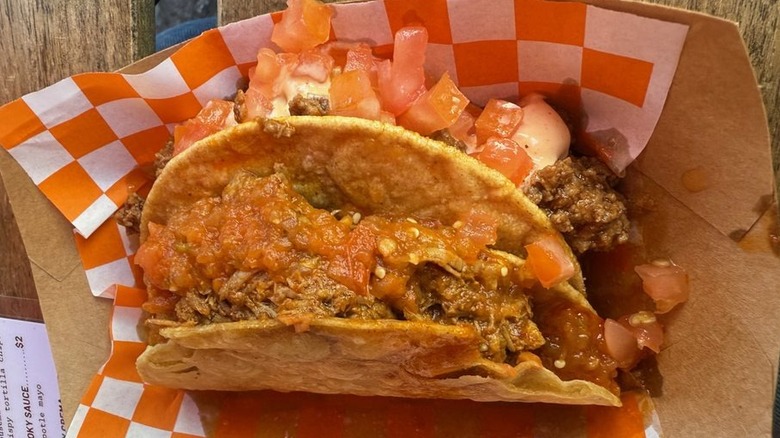A meat-filled hard shell taco in brown cardboard boat lined with red checkerboard paper; taco is covered with salsa and chopped tomatoes