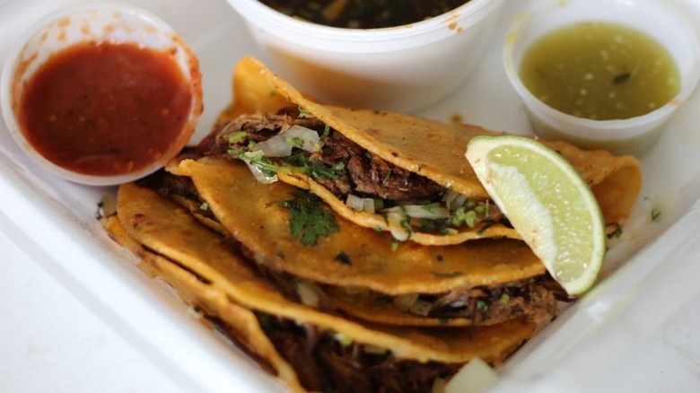 Three birria tacos tucked into the corner of a styrofoam to-go container with a ramekin of red sauce on the right, consumme in the center, and green sauce on the right; tacos garnished with a lime wedge