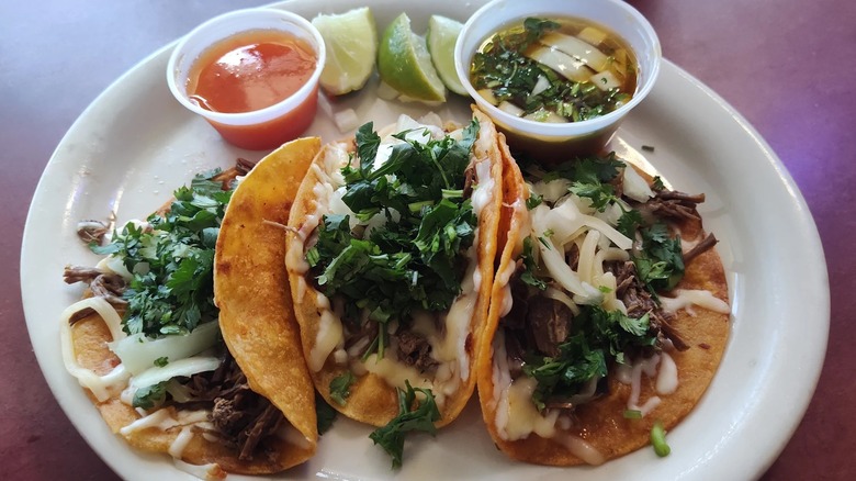 Three tacos in corn tortillas on a round white plate; each is filled with meat, white cheese, and cilantro; a plastic ramekin of red sauce is at top left, lime wedges in center, and a plastic ramekin of consumme is at the right