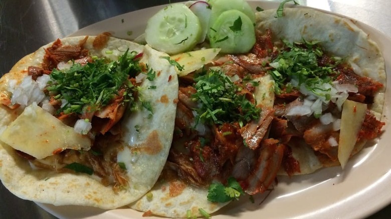 Three tacos on flour tortillas on an oval white plate; each is filled with meat and topped with chopped onions, cilantro, and shaved cheese; with a sliced cucumber/radish garnish