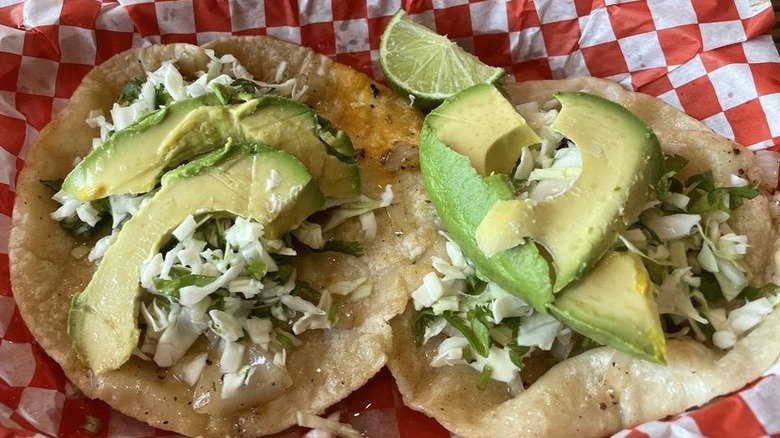 Two tacos on corn tortillas set in a red checkerboard-lined basket, each filled with sliced avocado atop a chopped mix of cabbage, onions, and cilantro; garnished with a lime wedge
