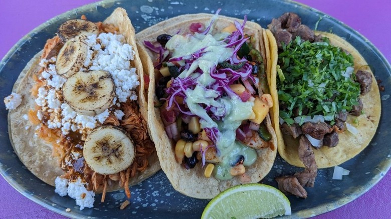 Three soft-shell tacos arranged on an oblong blue plate set on a purple background; left taco is filled with meat, crumbled white cheese, and sliced bananas; middle taco is filled with a mango-corn-black bean salsa topped with red cabbage and a creamy green sauce; right taco is filled with meat, chopped onion, and cilantro