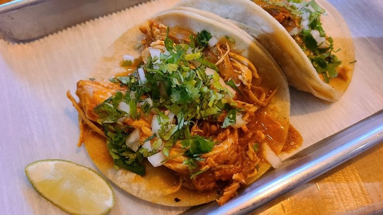 Two soft-shell tacos, filled with pulled chicken, chopped onions, and cilantro on a white paper-lined rectangular metal tray; one taco is flat at the center and the other is folded facing upper right; a lime wedge sits lower left