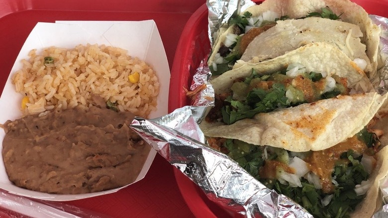 A cardboard boat of rice and beans on the left side of a red plastic tray with a red foil-lined basket with three tacos in it on the right; the corn tortillas are filled with cilantro and chopped onions covered in a brown sauce