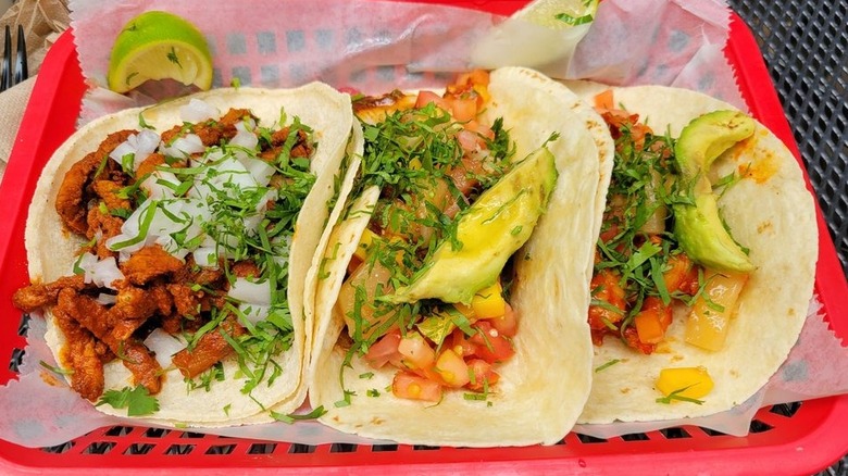 Three tacos in flour tortillas in a plastic red rectangular basket; taco on the right is filled with meat, chopped onions, and cilantro; the center and left tacos are filled with shrimp, chopped tomoatoes, an avocado slice, and cilantro