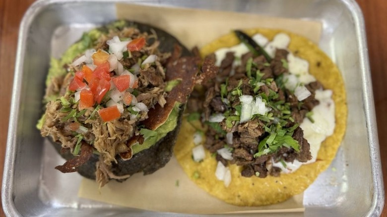 Two soft acos set on a beige-paper-lined metal tray; the left taco is a blue corn tortilla filled with guacamole, meat, and hipped tomatoes, onions, and cilantro; taco on the right is a yellow corn tortilla filled with white cheese, meat, and chopped onions and cilantro
