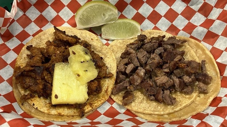 Two tacos on corn tortillas on red checkerboard paper; the left is filled with meat and two clises of pineapple; the right is filled with meat; two lime wedges sit at the top