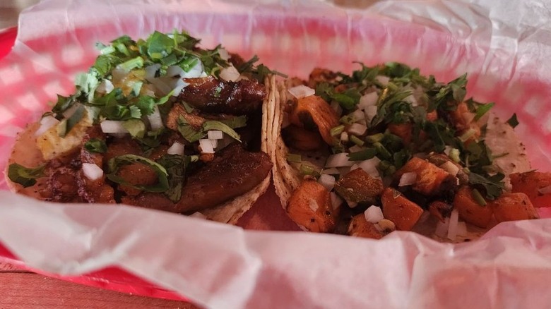 Two soft-shelled tacos in a wax-paper-lined oval-shaped red plastic basket; taco on the left is filled with meat, chopped onions, and cilantro; taco on the right is filled with cubed sweet potatoes, chopped onions, and cilantro