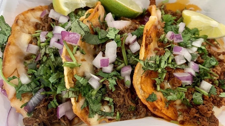A close-up of three tacos on fried corn tortillas filled with meat, chopped onions, and cilantro, garnished with lime wedges