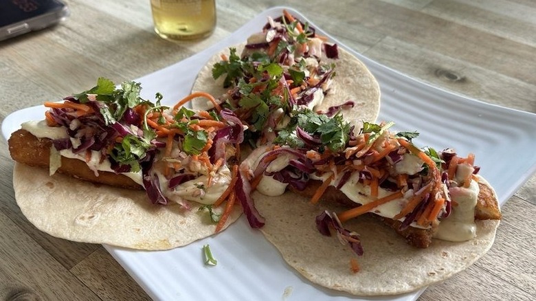 Three fried fish tacos topped with a white cream sauce and julienned carrots, cabbage, and cilantro, arranged on a square white plate