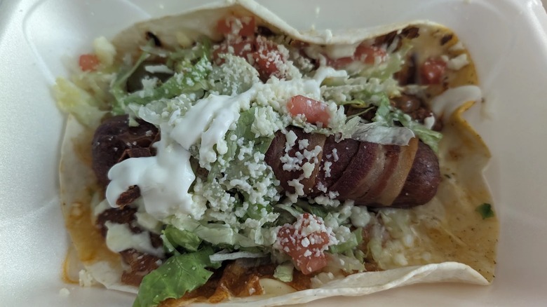 A bacon-wrapped hot dog topped with tomatoes, lettuce, cotija cheese, and sour cream in a flour tortilla