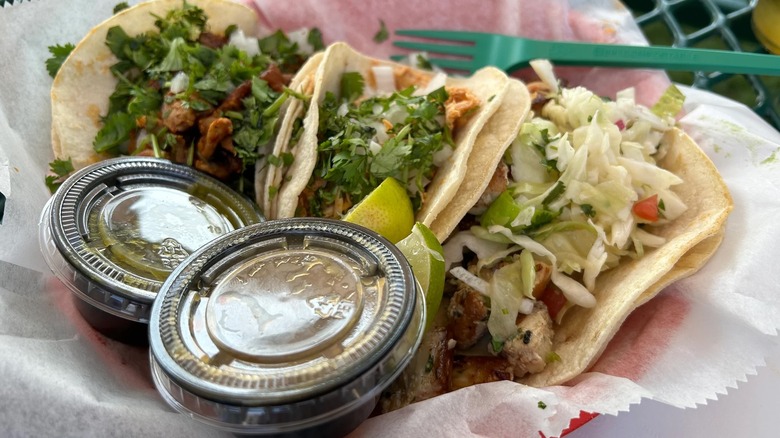 Three tacos in flour tortillas in a wax paper-lined red basket with two covered to-go ramekins of sauce in the foreground; the two left tacos are stuffed with meat and topped with cilantro; the right one is topped with shredded cabbage