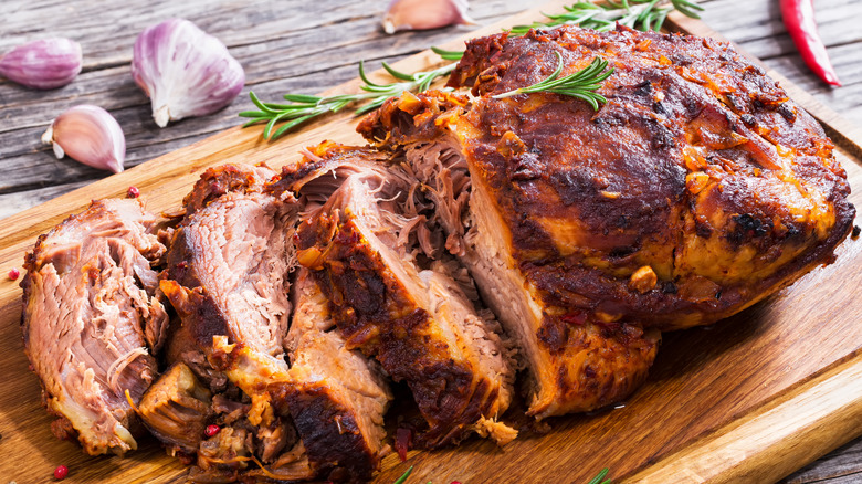 Big piece of slow cooked oven-barbecued pulled pork shoulder on chopping board with mixed peppercorns, rosemary, and garlic