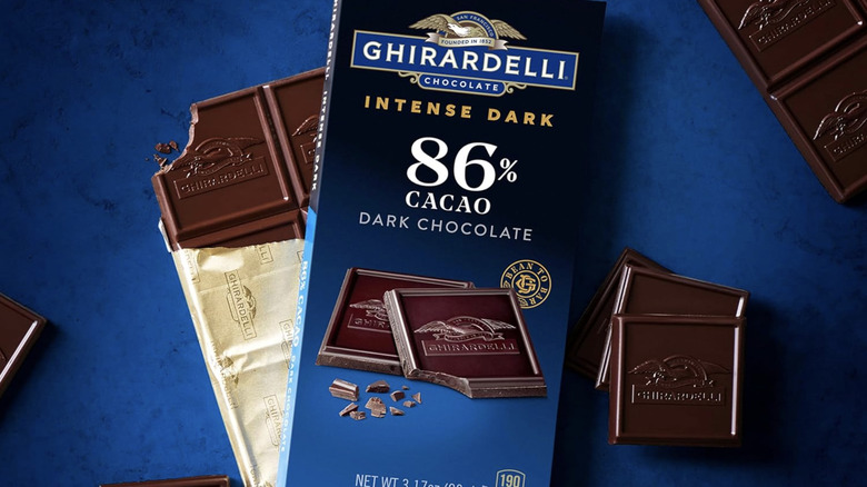 Unopened Ghirardelli Intense Dark Cacao Dark Chocolate Bar displayed over an opened bar with a bite mark on one corner, with both bars placed over a royal blue surface adorned with chocolate pieces.