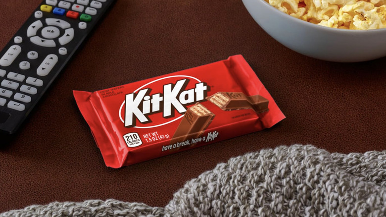 A KitKat bar placed over a wood surface alongside a remote control, a bowl of popcorn, and a knitted blanket.