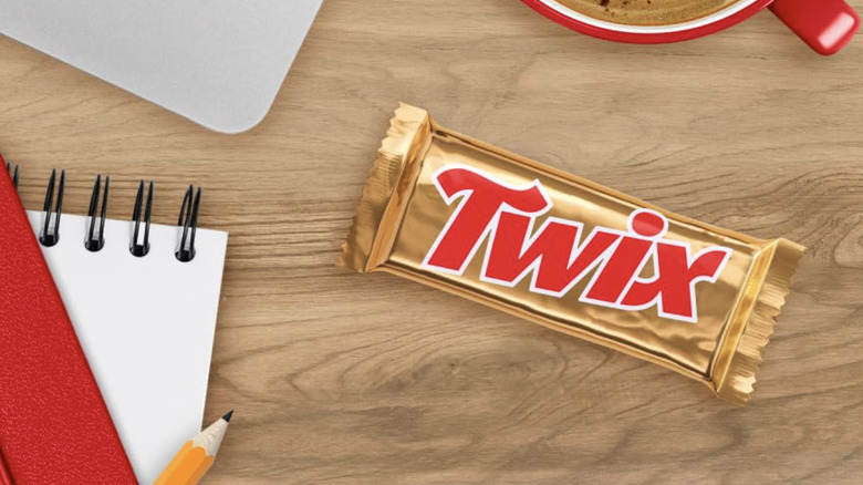 A Twix Chocolate Candy Bar placed on a wooden desk with many other items, including a laptop, coffee cup, notebook, and pencil.