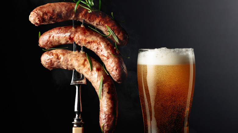 three grilled bratwursts on a fork next to a glass of beer