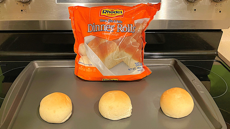 A bright orange bag of Rhodes frozen dinner rolls sit on a pan on a black stovetop with three baked rolls