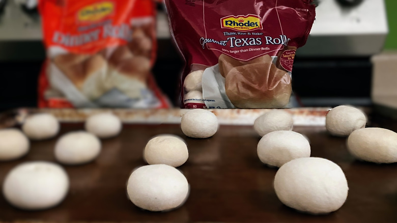 Two bags of Rhodes rolls on a stovetop with various sizes of frozen roll dough on a baking sheet