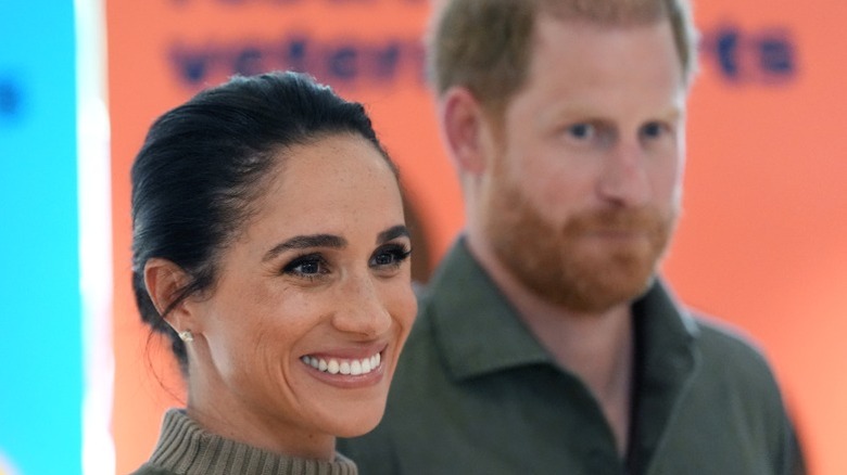 Princess Meghan at an event with Prince Harry in the background.