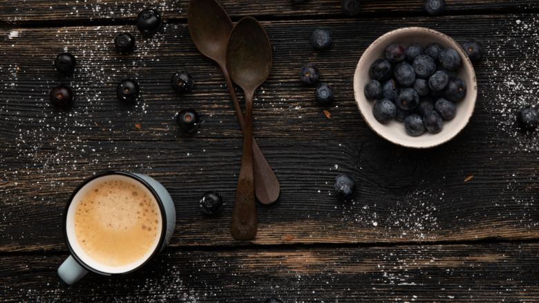 Overhead look at wooden table with two wooden spoons, blueberries scattered, with a white bowl filled blueberries, adjacent from a small white mug of coffee