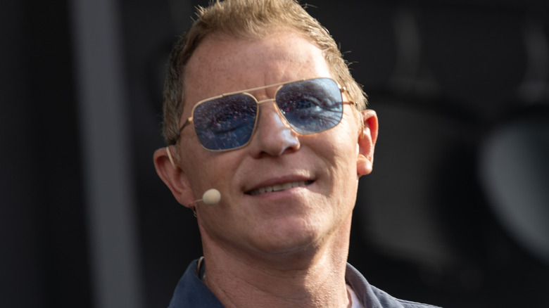 Bobby Flay wears blue sunglasses and a mic on stage at an event