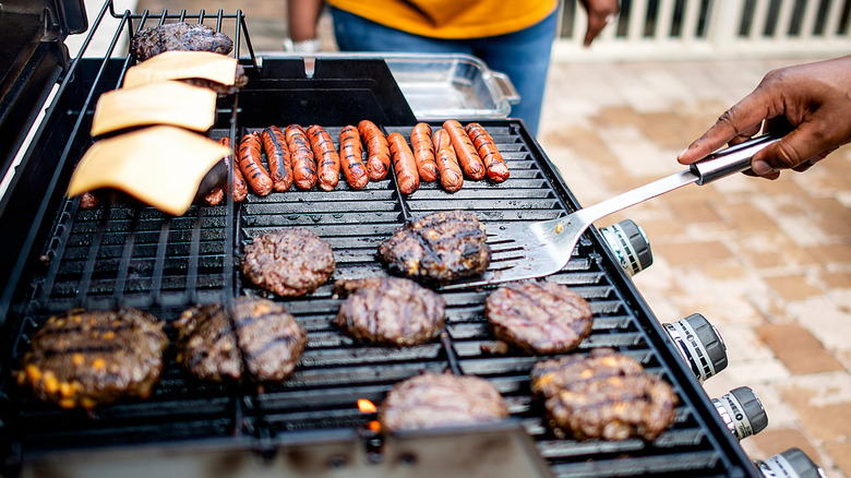 A person flipping burgers and cooking frankfurters on a grill.
