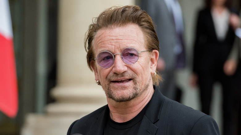 U2 singer Bono wearing round, purple-tinted glasses and a black T-shirt and jacket