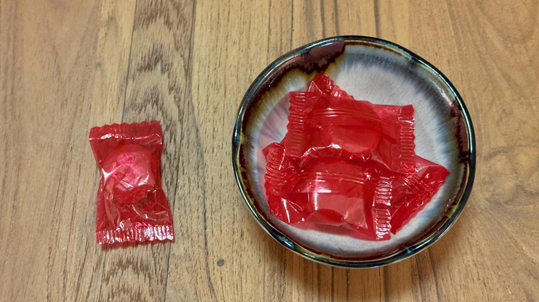 A small bowl of hard candy in red plastic wrappers, with one wrapped candy on the wooden table next to it