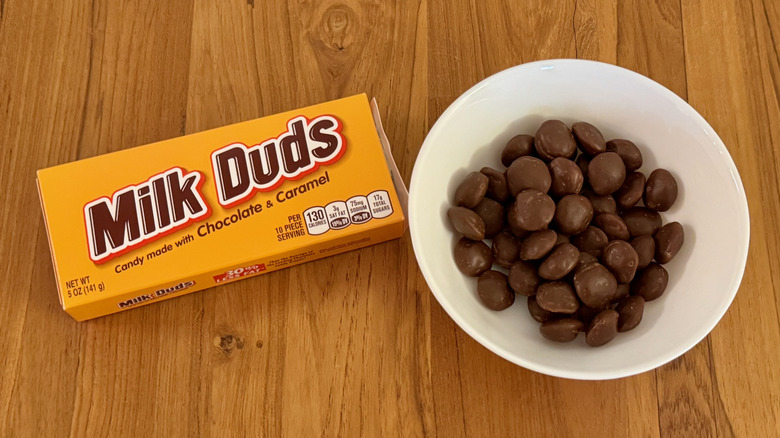 A white bowl filled with Milk Duds next to a yellow package on a wooden table.