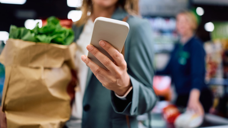 A woman is holding her phone in one hand with a bag of groceries in the other