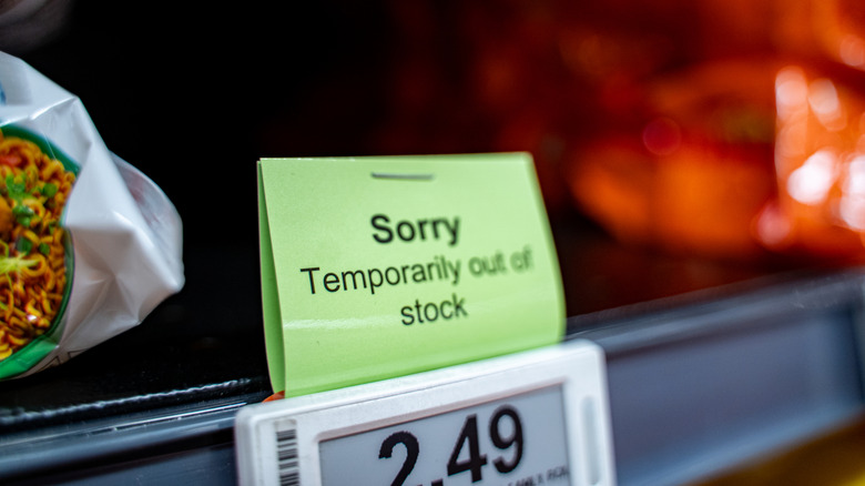A green temporarily out of stock sign is placed on a grocery shelf tag. There is empty space on the shelf behind it.
