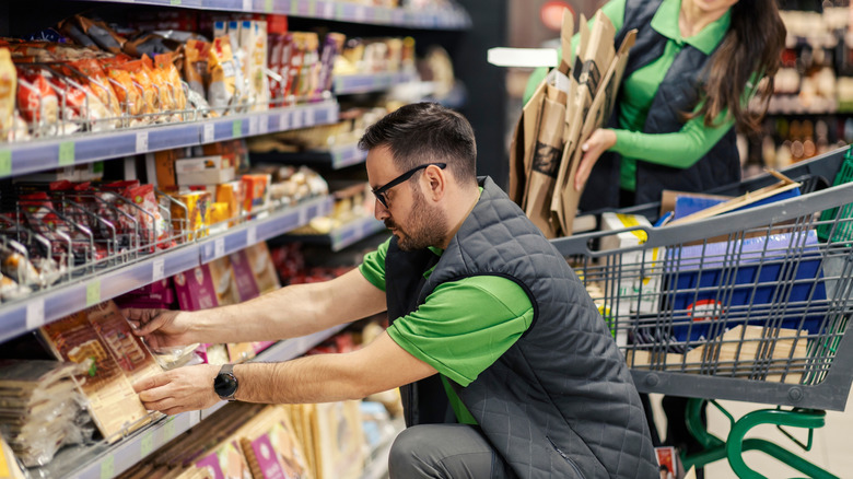 A grocery worker is knelt down, arranging a product on a shelf. A cart rests behind him, along with a coworker.