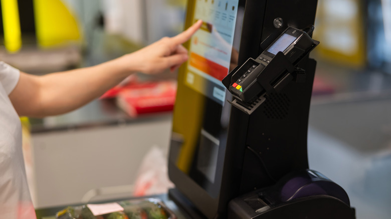 A picture of a self checkout counter, with a hand pressing a black button on the screen.