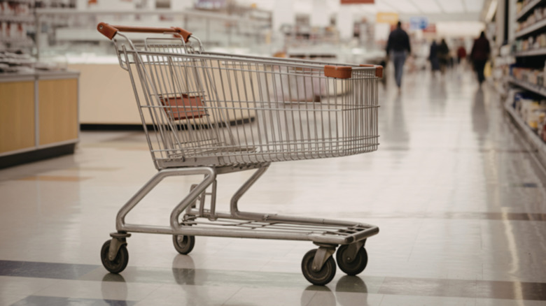 A lone shopping cart rests in one of the grocery store isles. People are walking in the background.