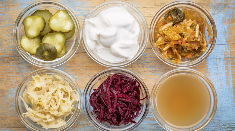 a set of fermented food great for gut health - top view of glass bowls against grunge wood: cucumber pickles, coconut milk yogurt, kimchi, sauerkraut, red beets, apple cider vinegar