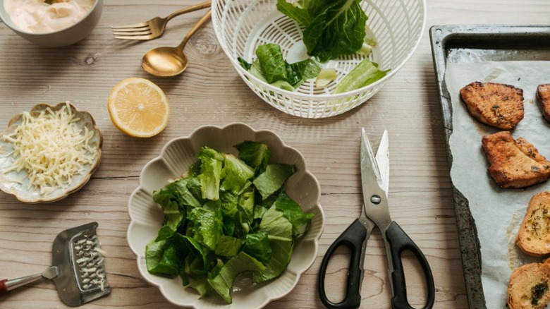 Ingredients for chicken Caesar salad on kitchen counter: bowl of lettuce, lemon, Parmesan cheese, scissors, utensils.