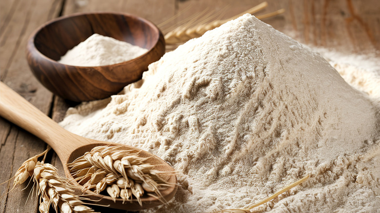 A mound of ground flour next to a wooden bowl and spoon with dried wheat